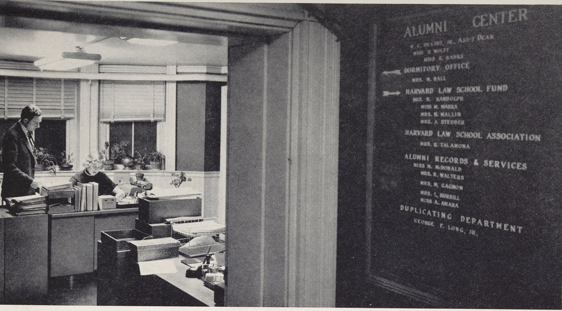 A view into an office. A man is standing next to a woman who is working on at a desk.